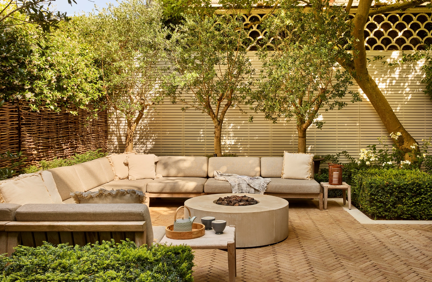 outdoor living room with c-shaped sofa, round fire pit coffee table, privacy screens, and trees