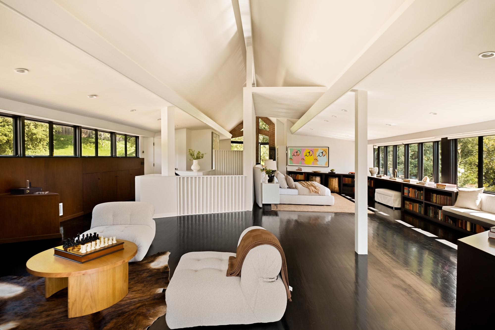 Image of an open concept living room and bedroom space. The floors are a black hardwood, the walls are full of windows, and the ceilings are angular and white with a few white beams in the space. There are two white chairs and a circular coffee table in the foreground. There is a bed and a tan rug in the background.
