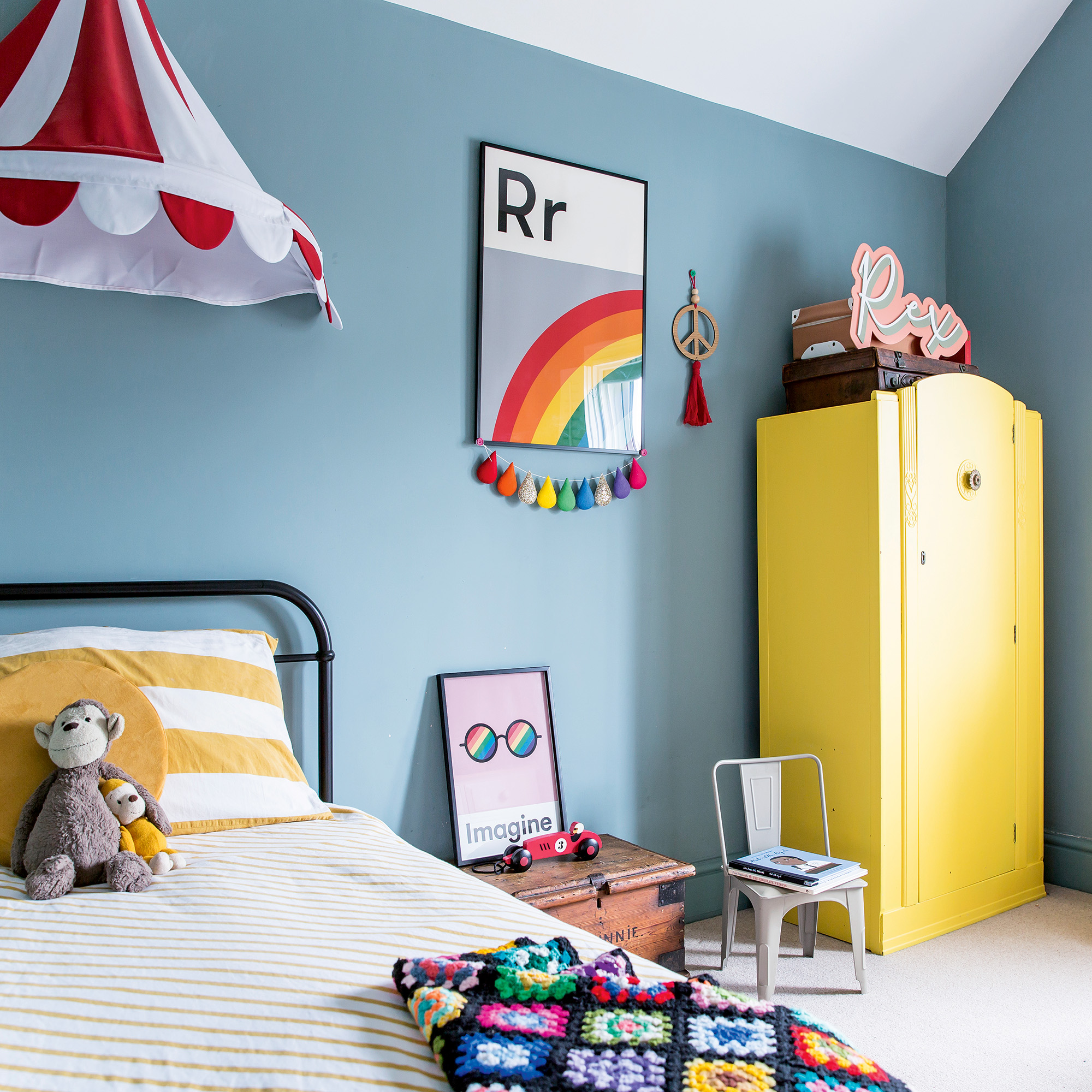 kids&#039; bedroom with blue walls, yellow wardrobe and cast iron bed with yellow and white striped bedlinen and two toy boxes