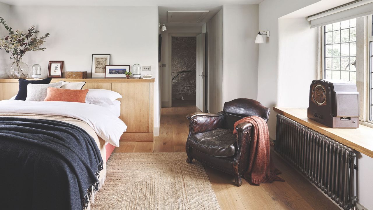 Large bedroom with wooden floor, jute rug, and textured bedding next to a leather armchair and black radiator