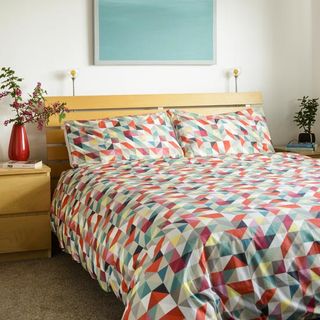 bedroom with white walls and brown carpet, a bed with a wooden slat bedhead and a colourful geometric duvet cover and a blue painting above the bed