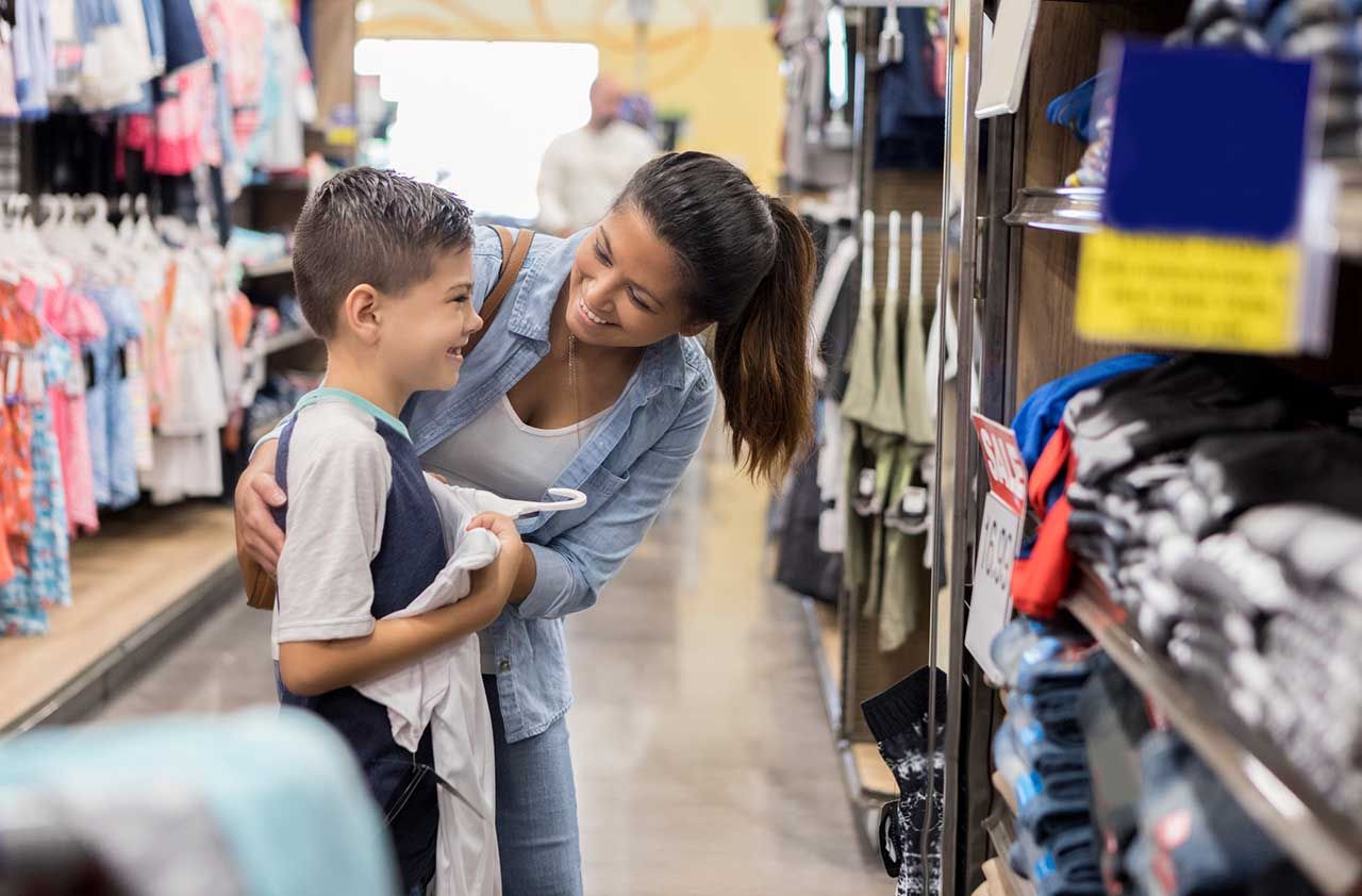 Happy young boy looks at a shirt in the mirror while shopping for back to school clothes with his mom.