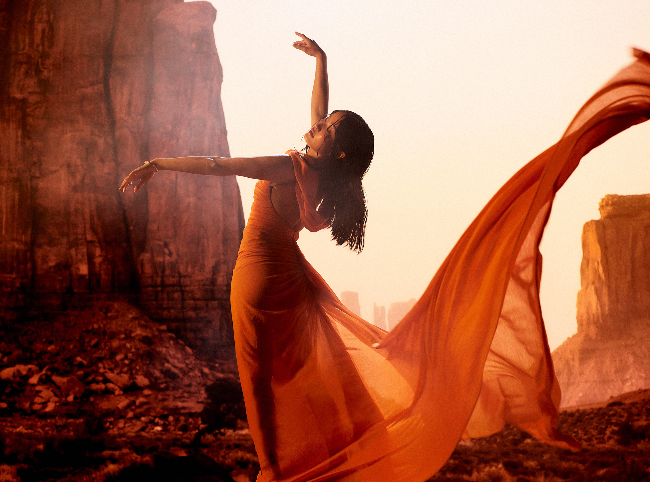 Anna Sawai poses in a flowing orange dress against a sunlit canyon landscape.