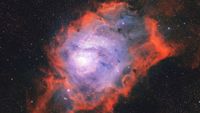 A nebula is photographed glowing in deep space among a sea of stars. It features a blue glowing core reminiscent of a lagoon, surrounded by red, flame-like clouds.