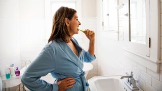 Woman brushing teeth in bathroom mirror, wearing blue robe
