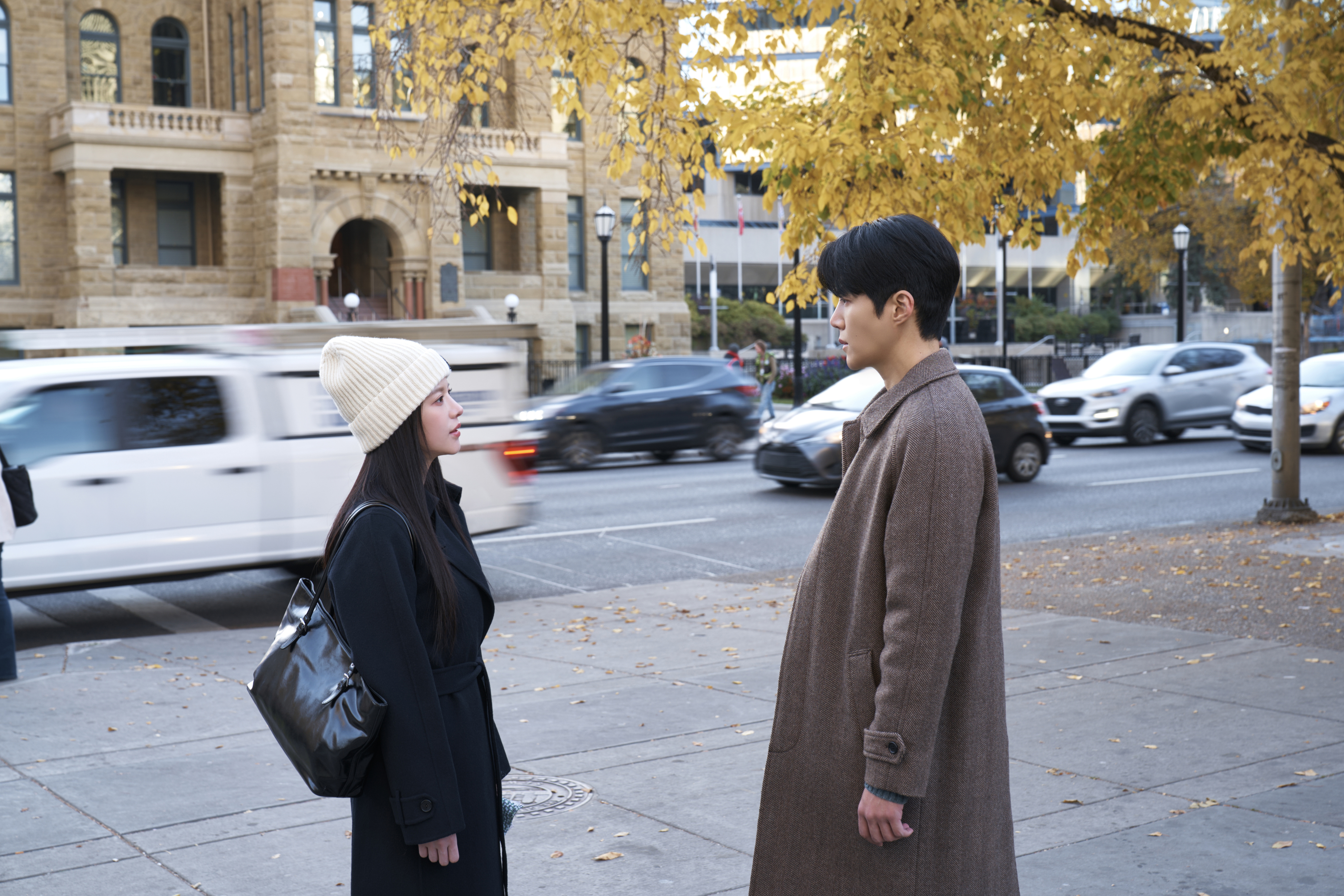 A woman in a white beanie (Go Youn-jung) and a man in a brown coat (Kim Seon-ho) face each other while standing on a city sidewalk. Cars are slightly blurred as they move behind them, with a tree with fall leaves and a stone building in the background, in the Netflix K-drama 'Can This Love Be Translated?'