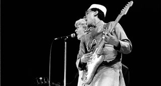 A black and white action shot of David Bowie and Carlos Alomar sharing the mic onstage, with Alomar playing his Fender Stratocaster