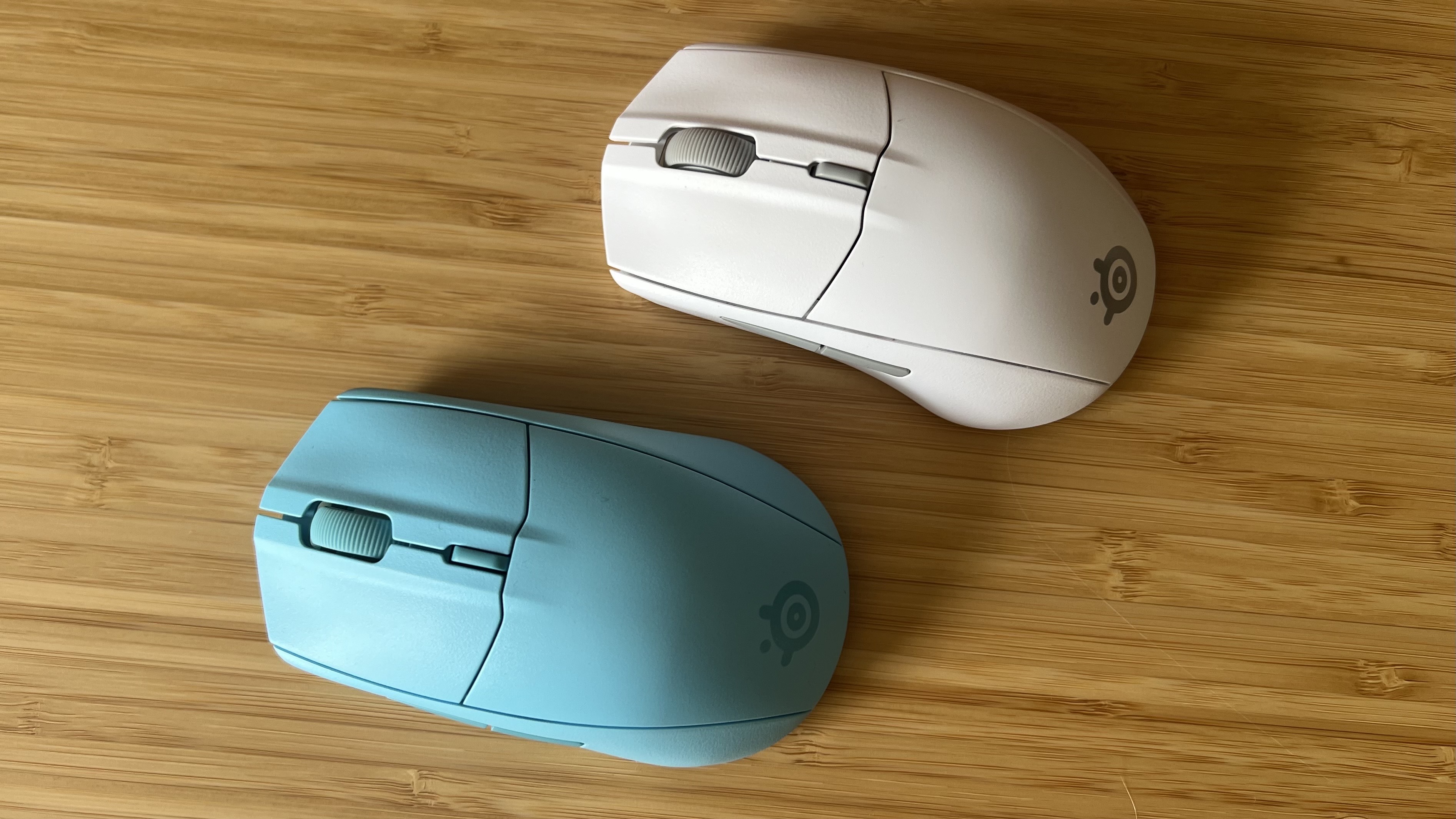 Topo down shot of two SteelSeries Rival 3 Wireless Gen 2 gaming mice, one blue and one white, on a wooden desk