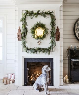 A fireplace with foliage garland and 'Joy' neon sign with dog sitting below