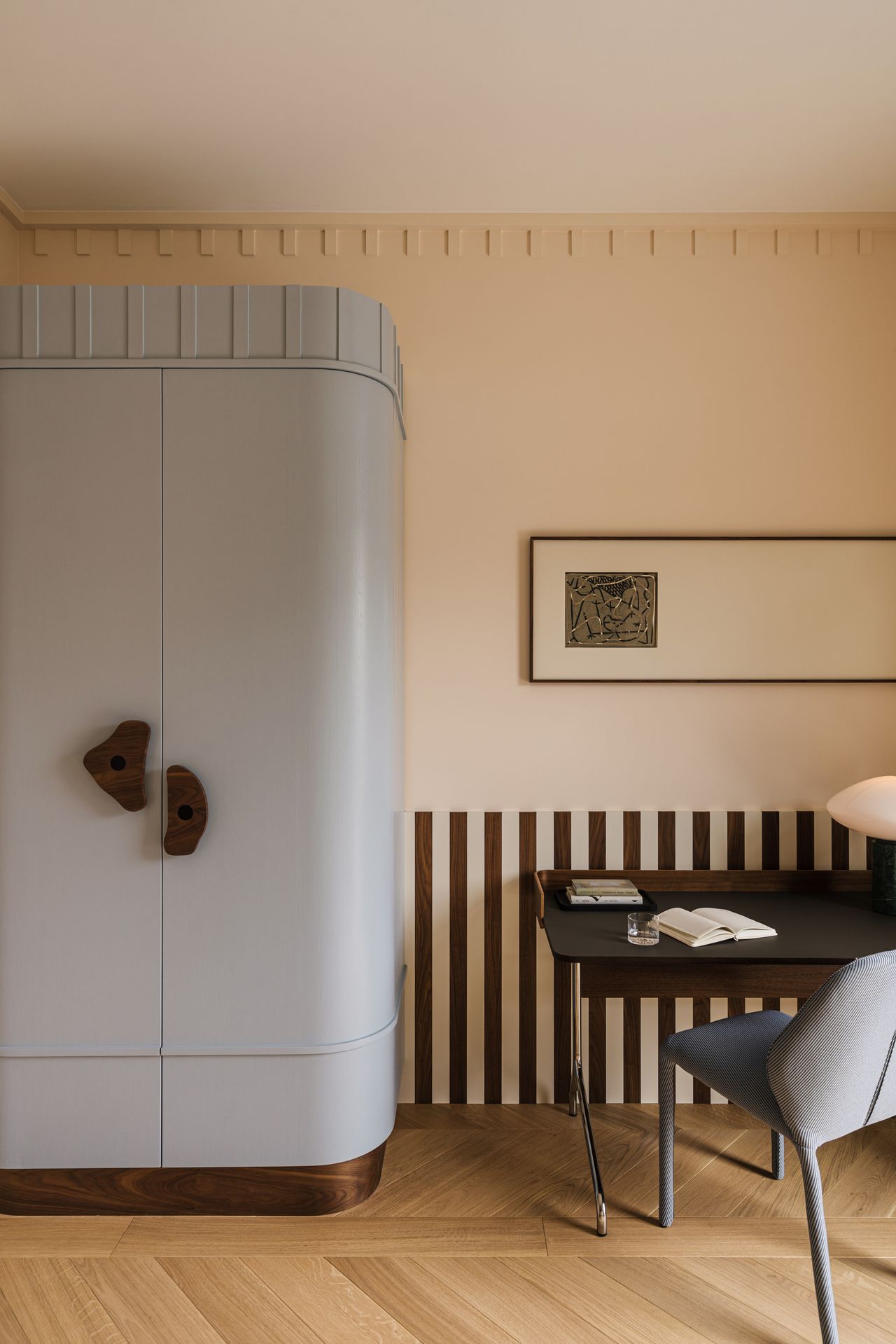 Image of a pinkish-beige room with the bottom half of the wall in brown stripes. There is a large light blue wardrobe with brown knobs and a brown base. There is a brown desk with a light blue desk chair.