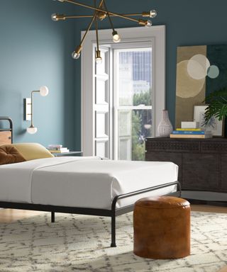 A blue bedroom with a white bed with a metal frame, a rug, brown ottoman, and a black set of drawers