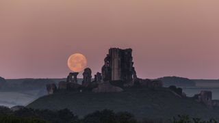 Full moon rising behind Corfe Castle, Dorset, U.K. A castle ruin is in the foreground of the image with a subtle pink hued sky and a bright full moon. 