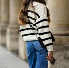 A passerby wears a white with black striped oversized pullover, a gold ring, blue denim large jeans pants, a black shiny leather crossbody bag, on November 05, 2021 in Paris, France. (Photo by Edward Berthelot/Getty Images)