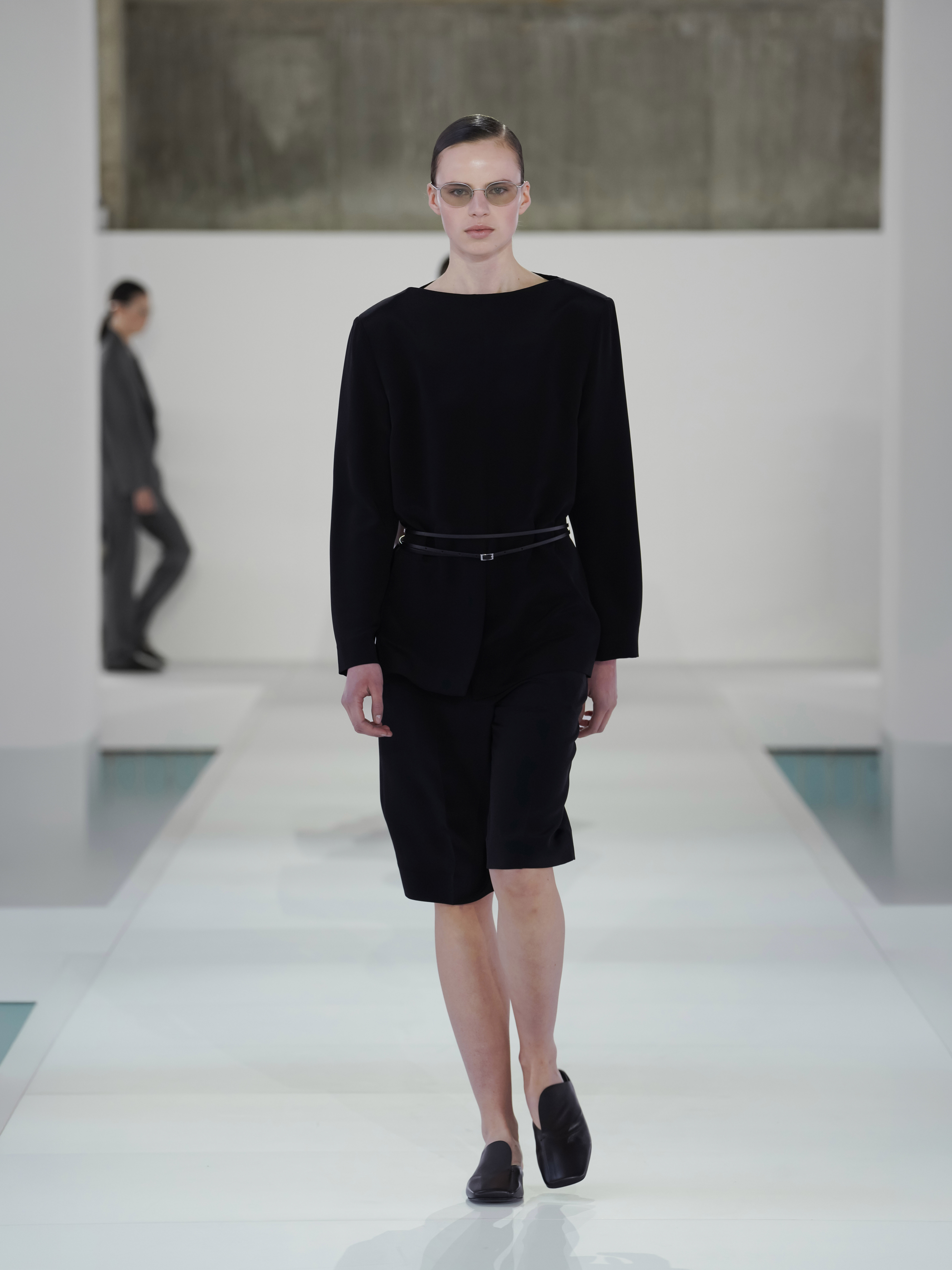 A model walking in the SS26 COS show in Seoul, South Korea, wearing a black collarless jacket and Bermuda shorts.