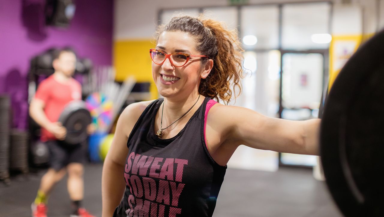 Woman in gym leans on a barbell in a squat rack. Her hair is in a pony tail and she is wearing red glasses and a black tank top with the words &quot;sweat today smile&quot; visible