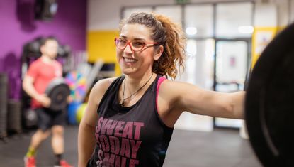 Woman in gym leans on a barbell in a squat rack. Her hair is in a pony tail and she is wearing red glasses and a black tank top with the words "sweat today smile" visible