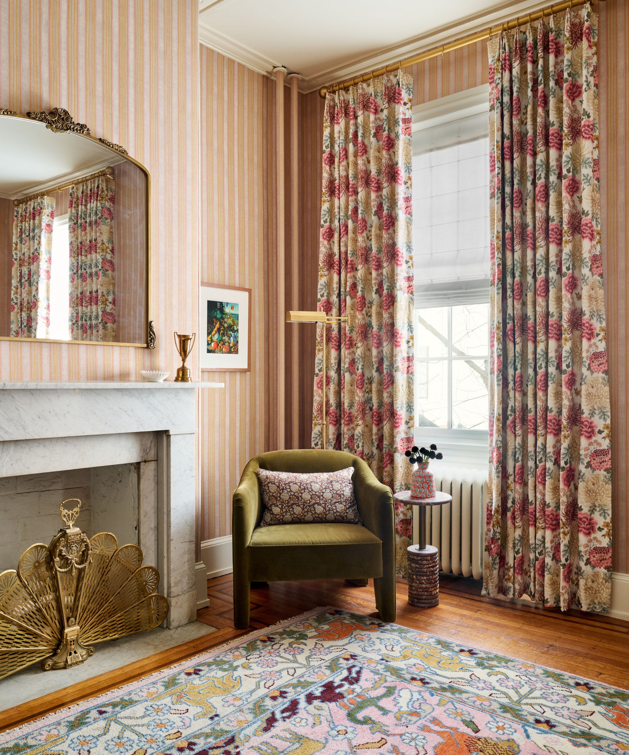 Bedroom corner with a marble fireplace, gold arch mirror, moss-green velvet armchair, and bold pink floral curtains.