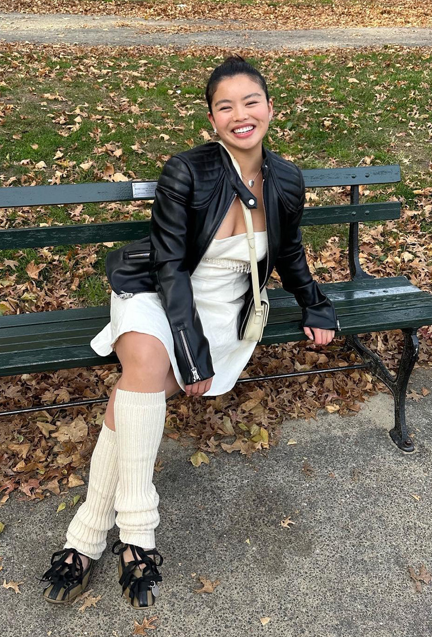 The best ballet sneakers are shown in a photo of a woman sitting on a bench in the park wearing a black leather moto jacket with a white mini dress, white leg warmers, and olive green ballerina sneakers