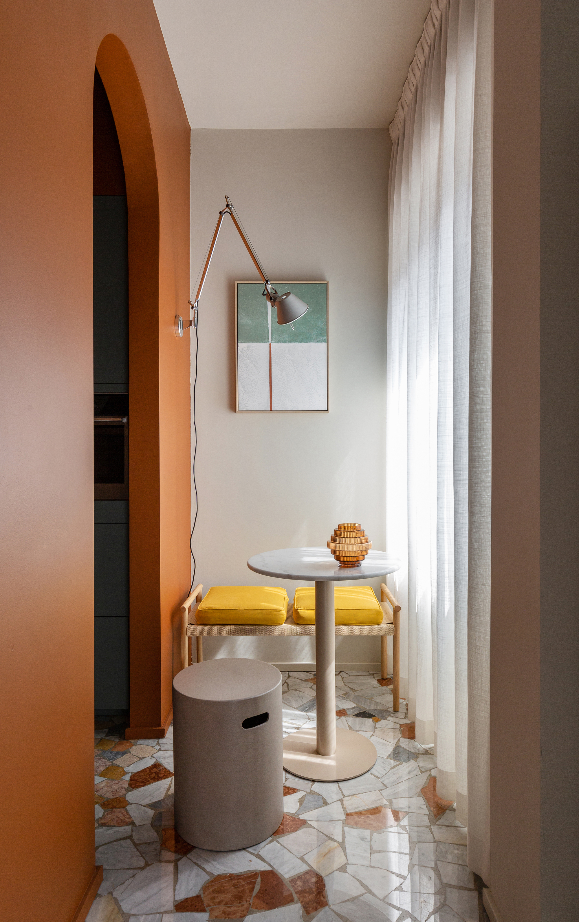 Image of a hallway where the walls are painted a warm white and a bright, dark orange. There is a bench seat with a yellow cushion at the far end and a small round metal table and matching stool. The floors are a white and orange Palladiana terrazzo tile.