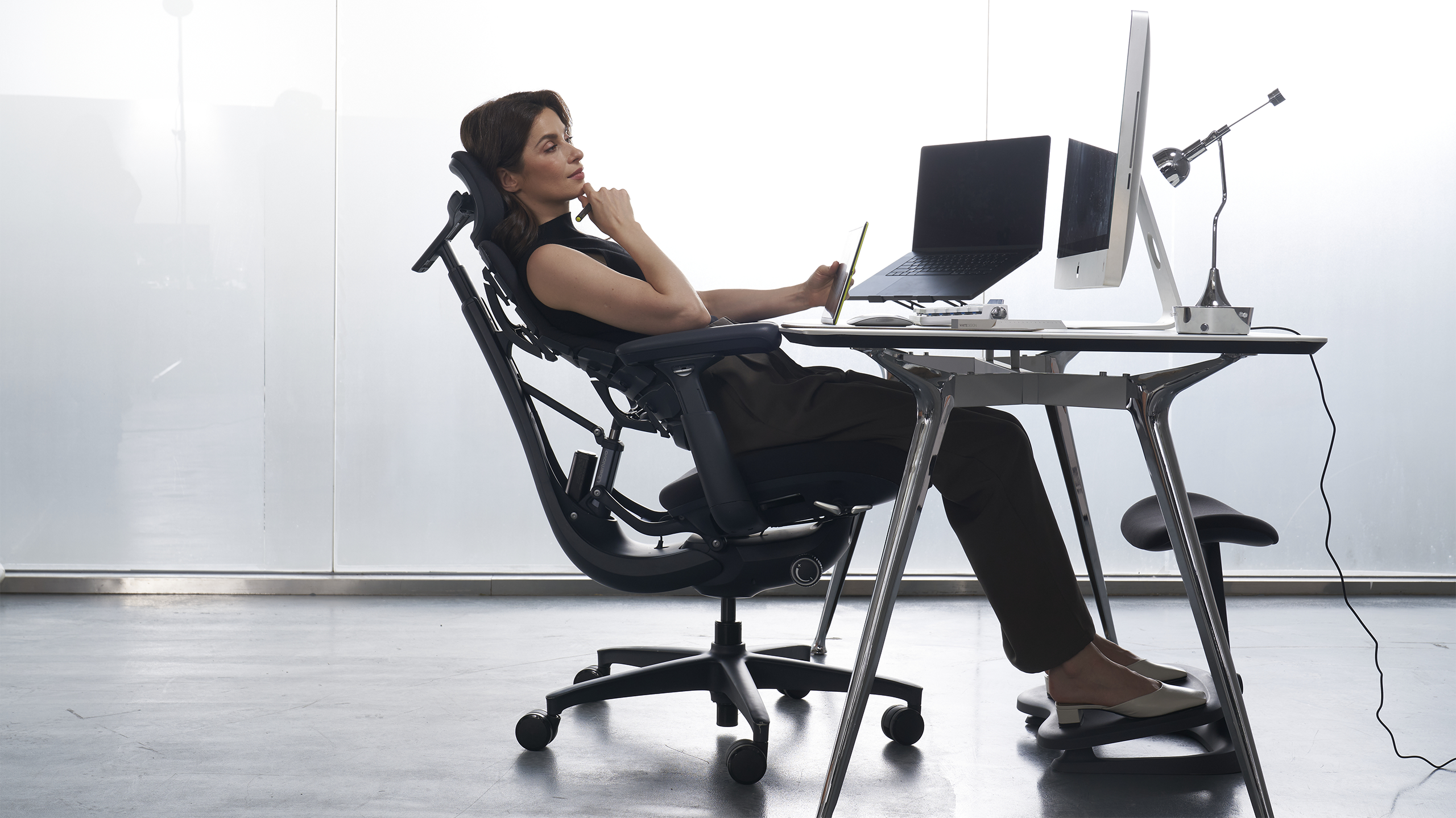A woman sits back in a sleek ergonomic office chair at a modern desk, holding a smartphone while facing a desktop computer and open laptop in a bright, minimalist workspace with frosted glass walls.
