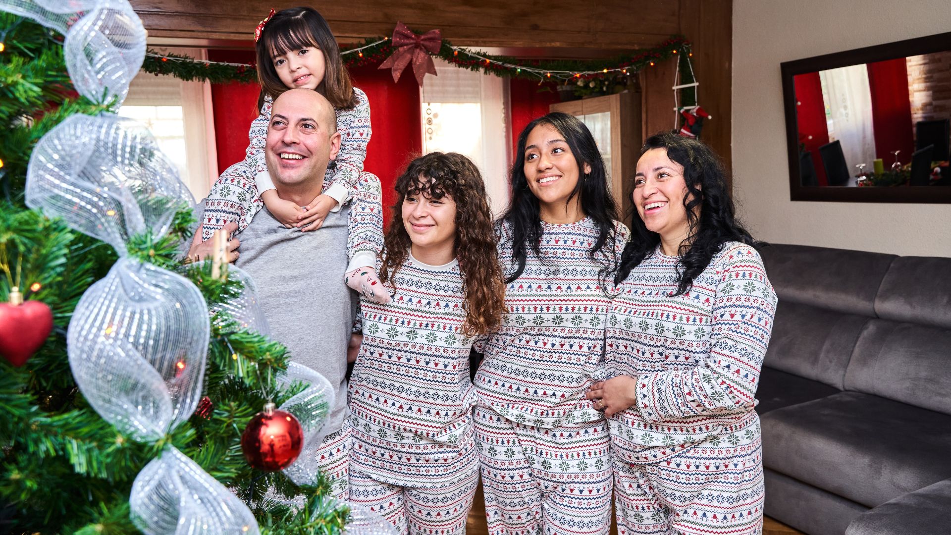 A young family wearing matching Christmas-themed pajamas beam at a beautiful Christmas tree in their modern living room.