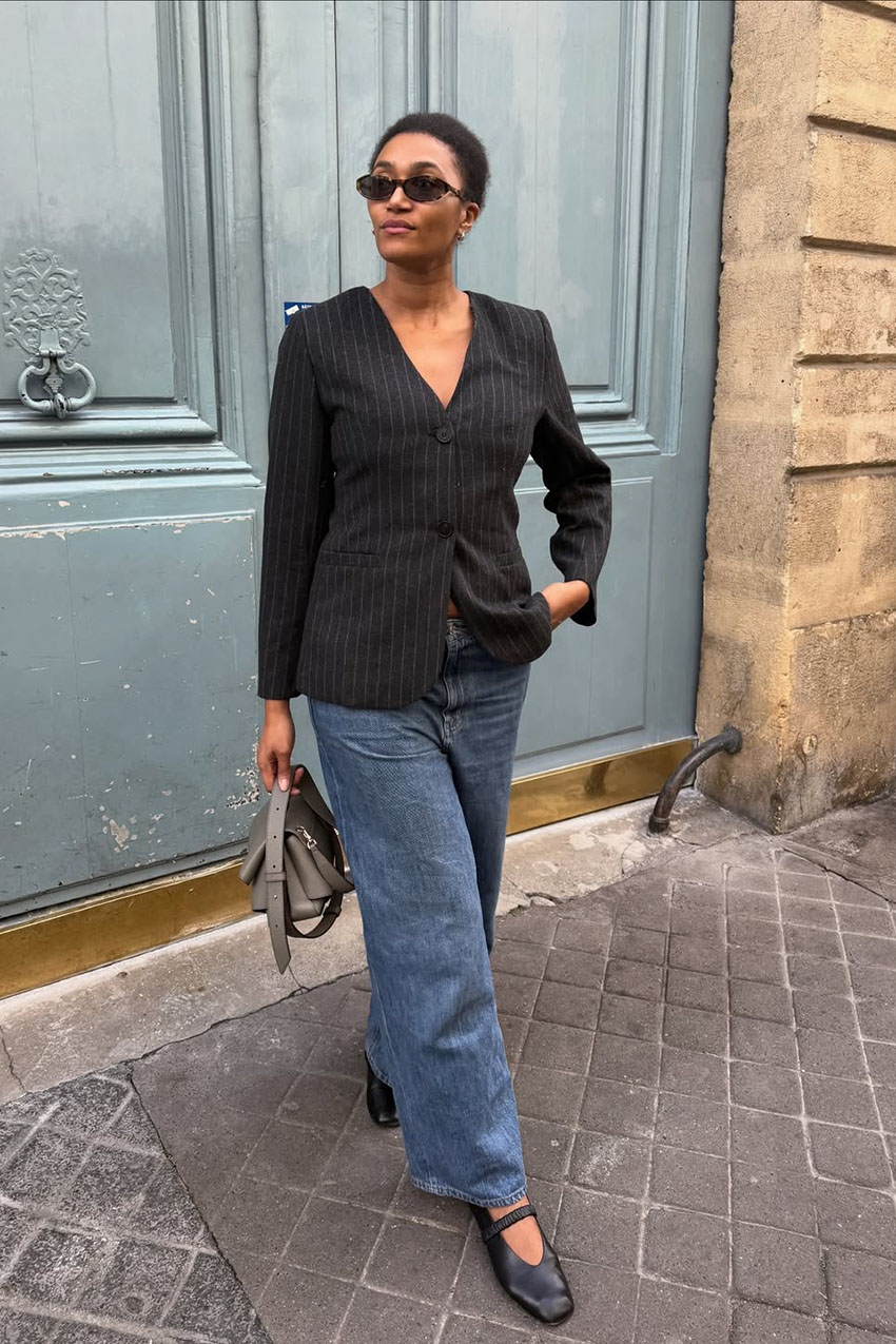 A woman wearing a black blazer, blue straight-leg jeans, black flats, and sunglasses.