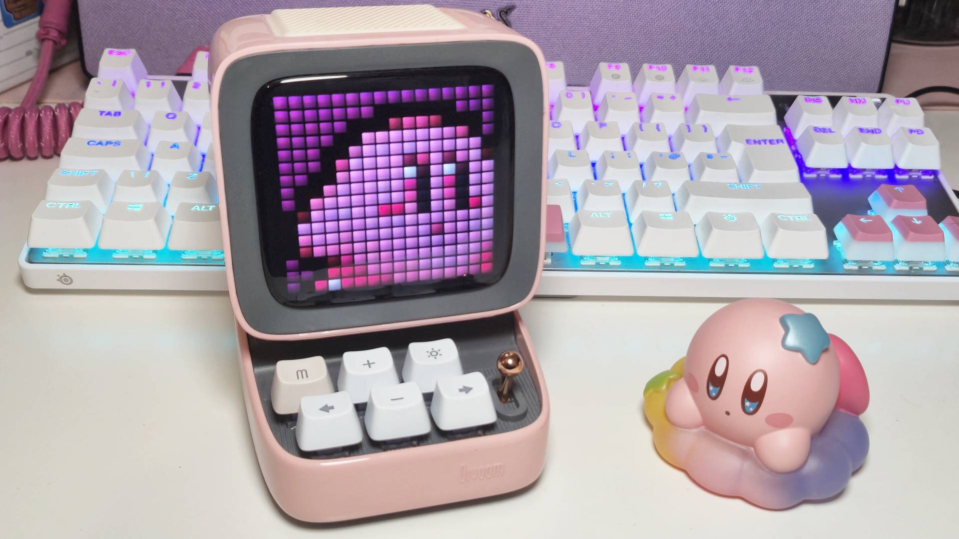 Photo by Rosalie Newcombe of the Divoom Ditoo speaker with art by Divoom user Twilights on the display, and a Kirby toy by Bandai sitting next to it.