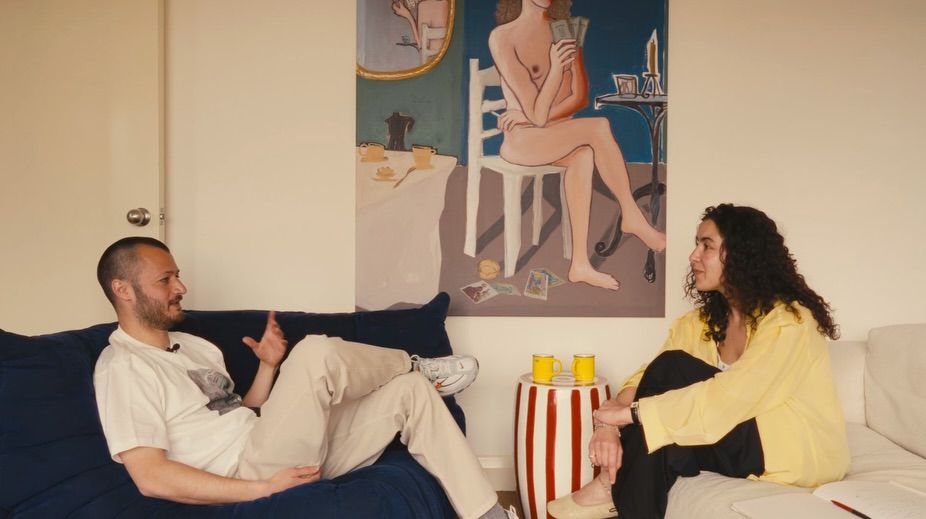 The interior of a young, brunette, curly-haired woman's living room-cum-kitchen, designed with pops of unexpected color, unique artwork, and sculptural sofas in blue and cream.