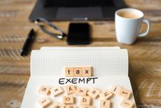 Closeup on notebook over vintage desk surface, front focus on wooden blocks with letters making Tax Exempt text with office tools and coffee cup in background