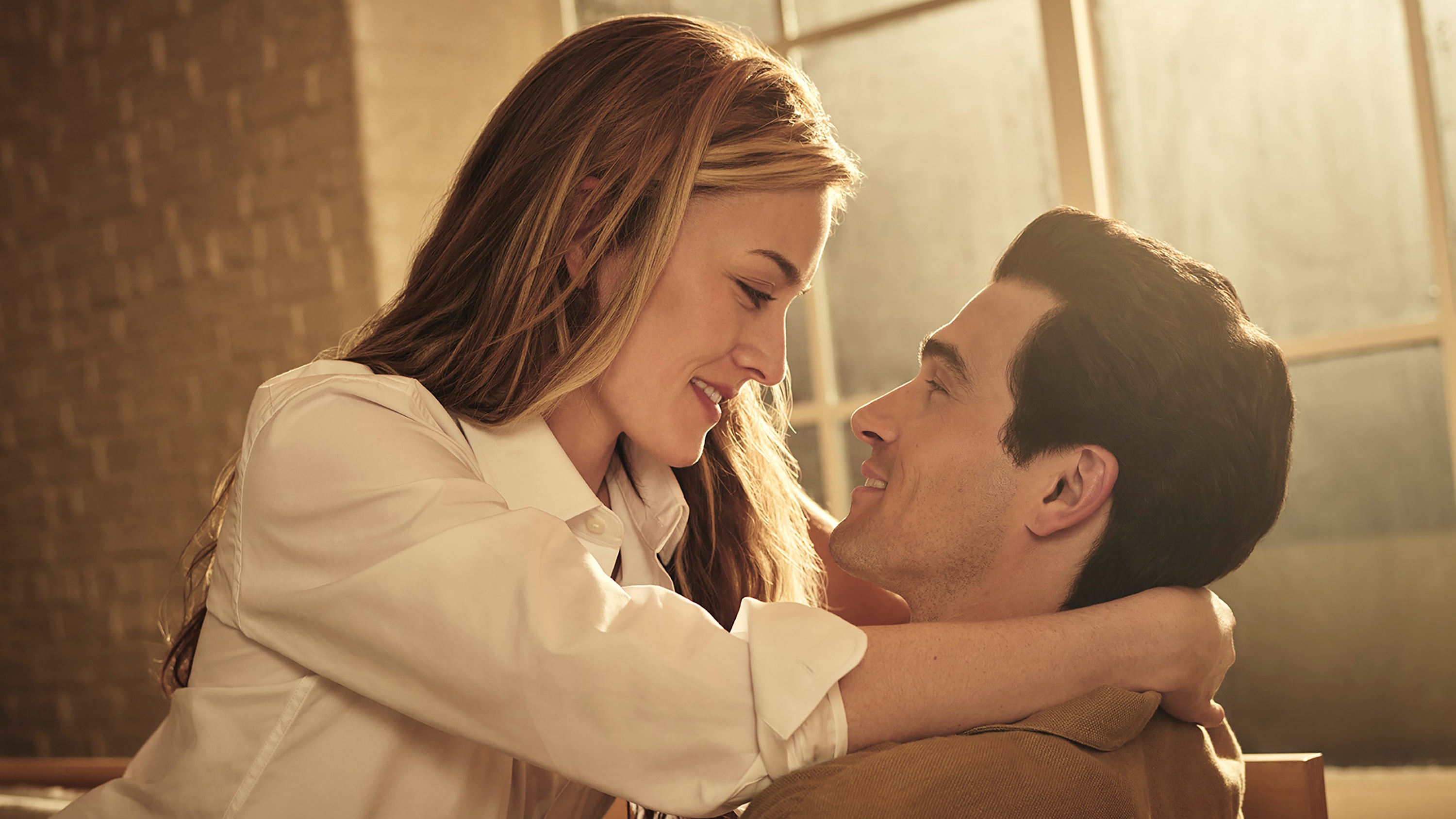 Sarah Pidgeon as Carolyn Bessette Kennedy and Paul Kelly as John F. Kennedy Jr. Carolyn and John smile at each other as she sits on his lap with her arms around his neck.