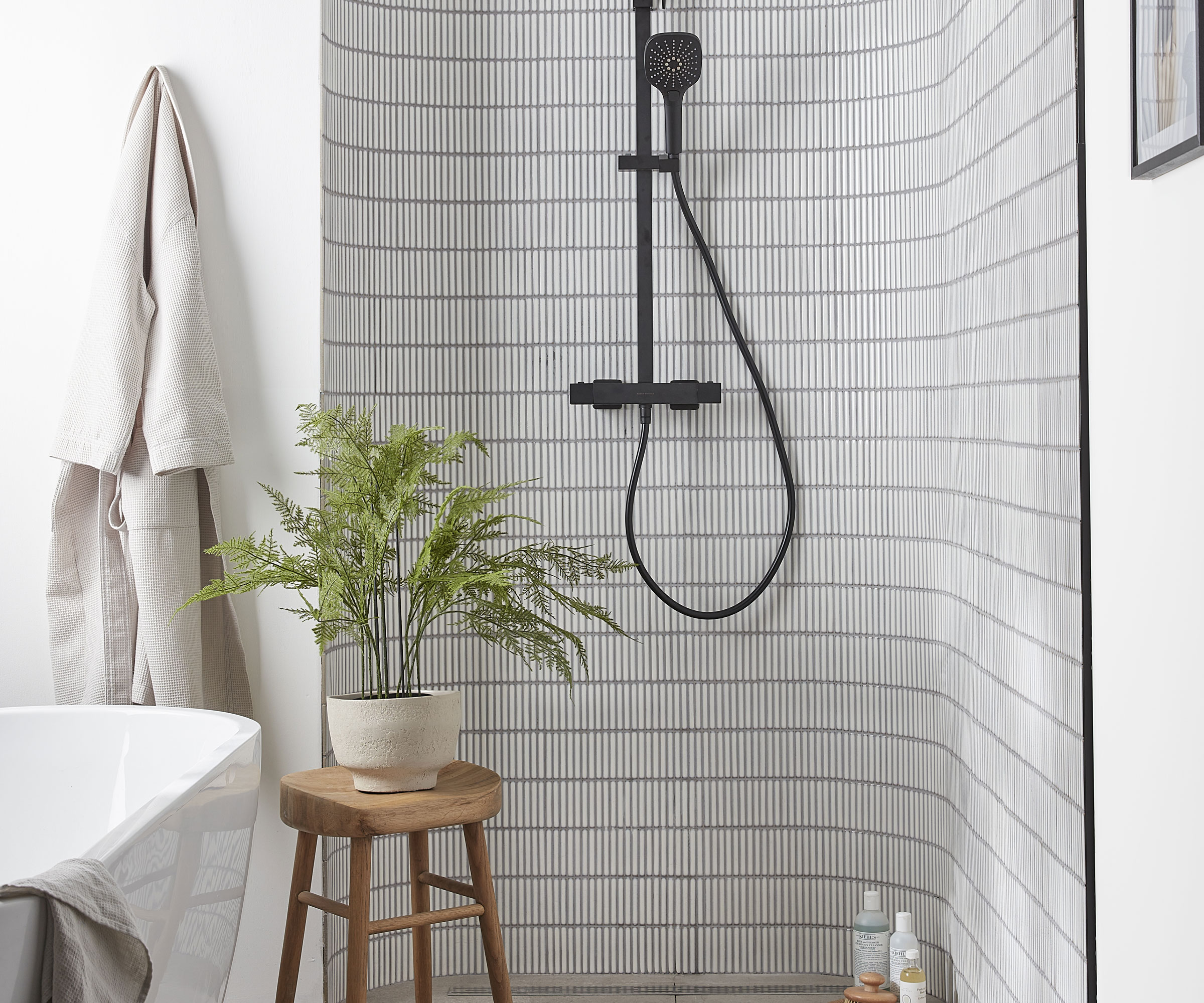 Thin white vertical linear mosaic tiles on shower wall with black shower and wooden stool and plant to side next to free standing bath