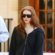 Sadie Sink wearing a black zip-up hoodie, low-rise long denim shorts, and black leather flats