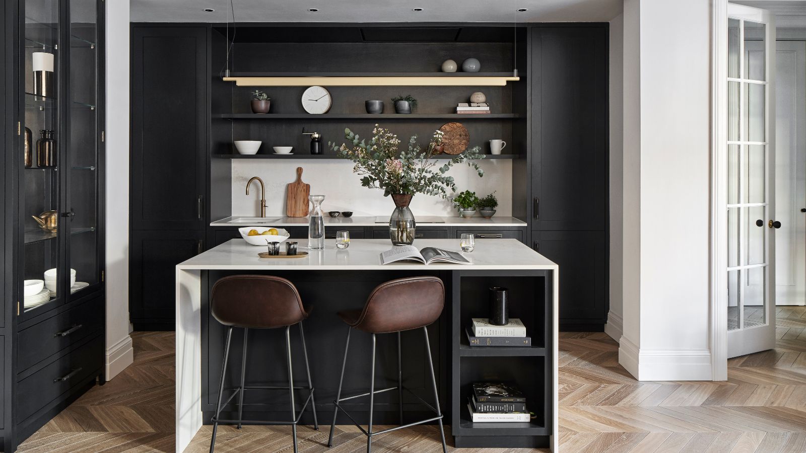 A black kitchen with wooden flooring, a white island with brown bar stools, and black shelving filled with bowls, chopping boards and a white clock. To the right is a glass and black cabinet and to the right is a white wall and white glass paned open door.