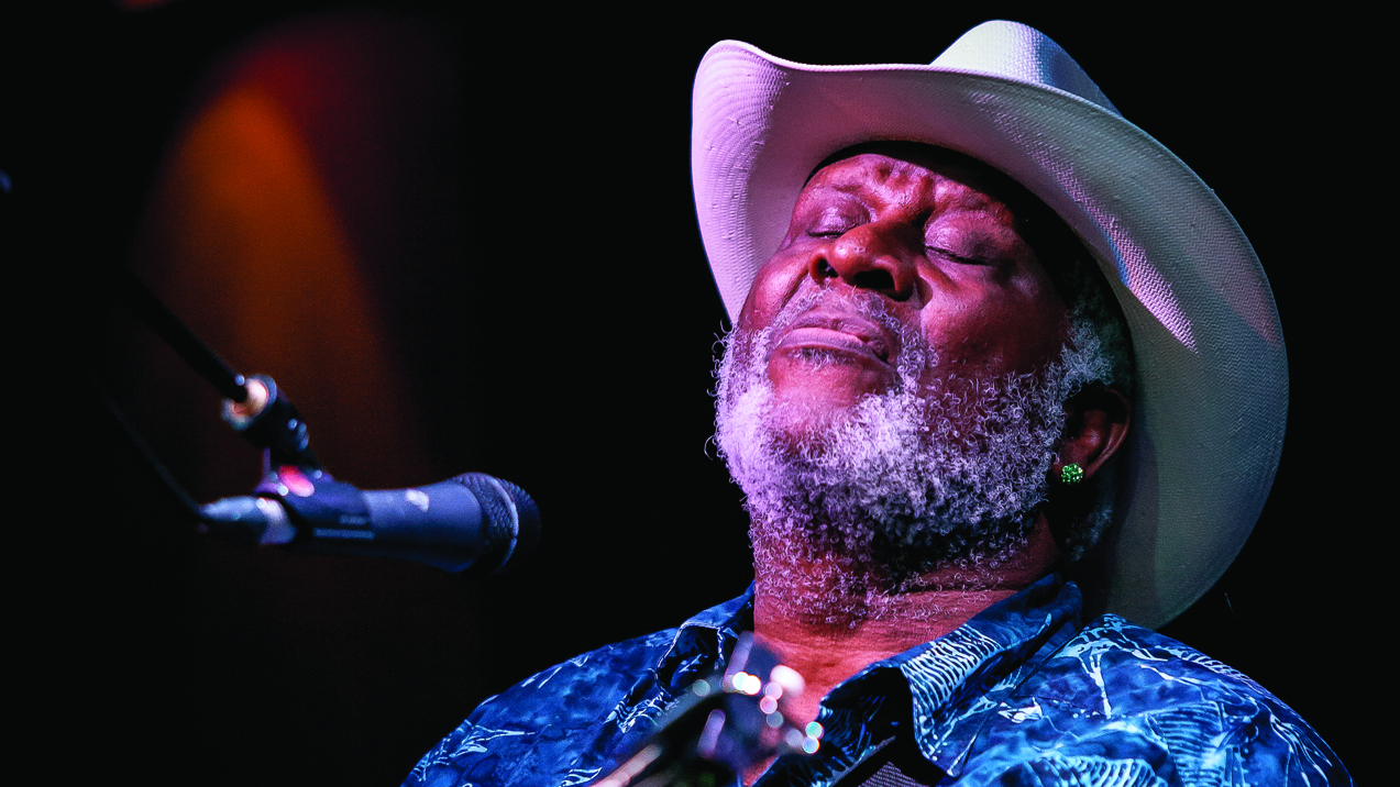 Taj Mahal onstage, in a white hat, with his eyes closed.