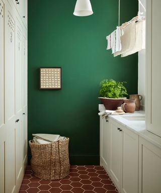 small laundry room with hexagon floor tiles, off white cabinetry, laundry maid, green wall, basket full of linen, sink, pendant light, ceramics