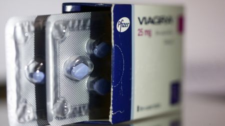 Close-up image of an opened box of blue, diamond-shaped Viagra tablets with two pill packets sticking out towards the camera. The Pfizer logo can be clearly seen on the side of the box.