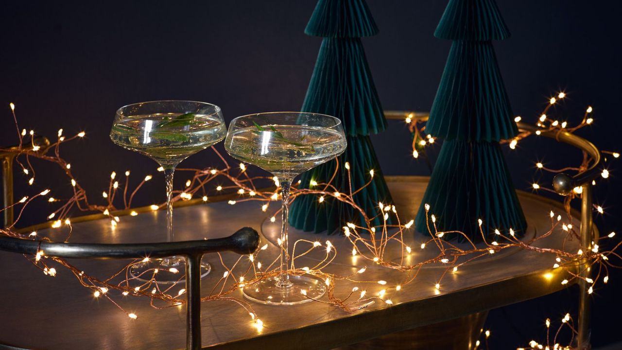 A string of small fairy lights draped across a bronze bar cart with two paper trees and two drinking glasses filled with champagne in the center