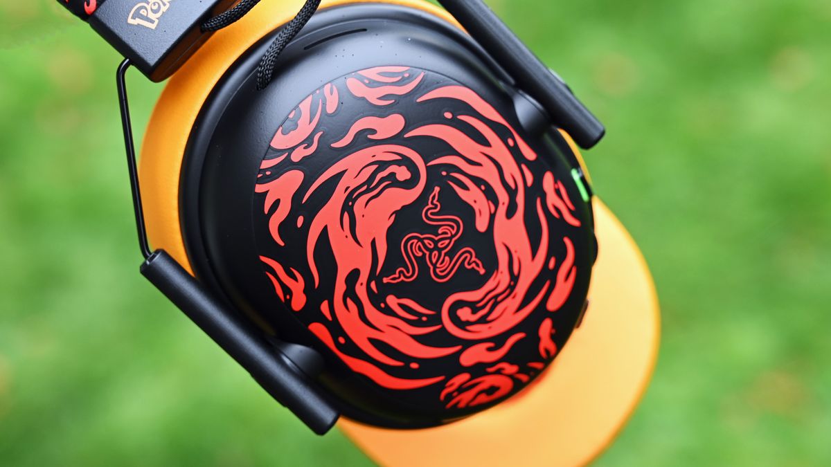 Close-up of the black earcup faceplate featuring swirling red flame designs around the Razer logo, framed by a yellow cushion and metal wire slider.