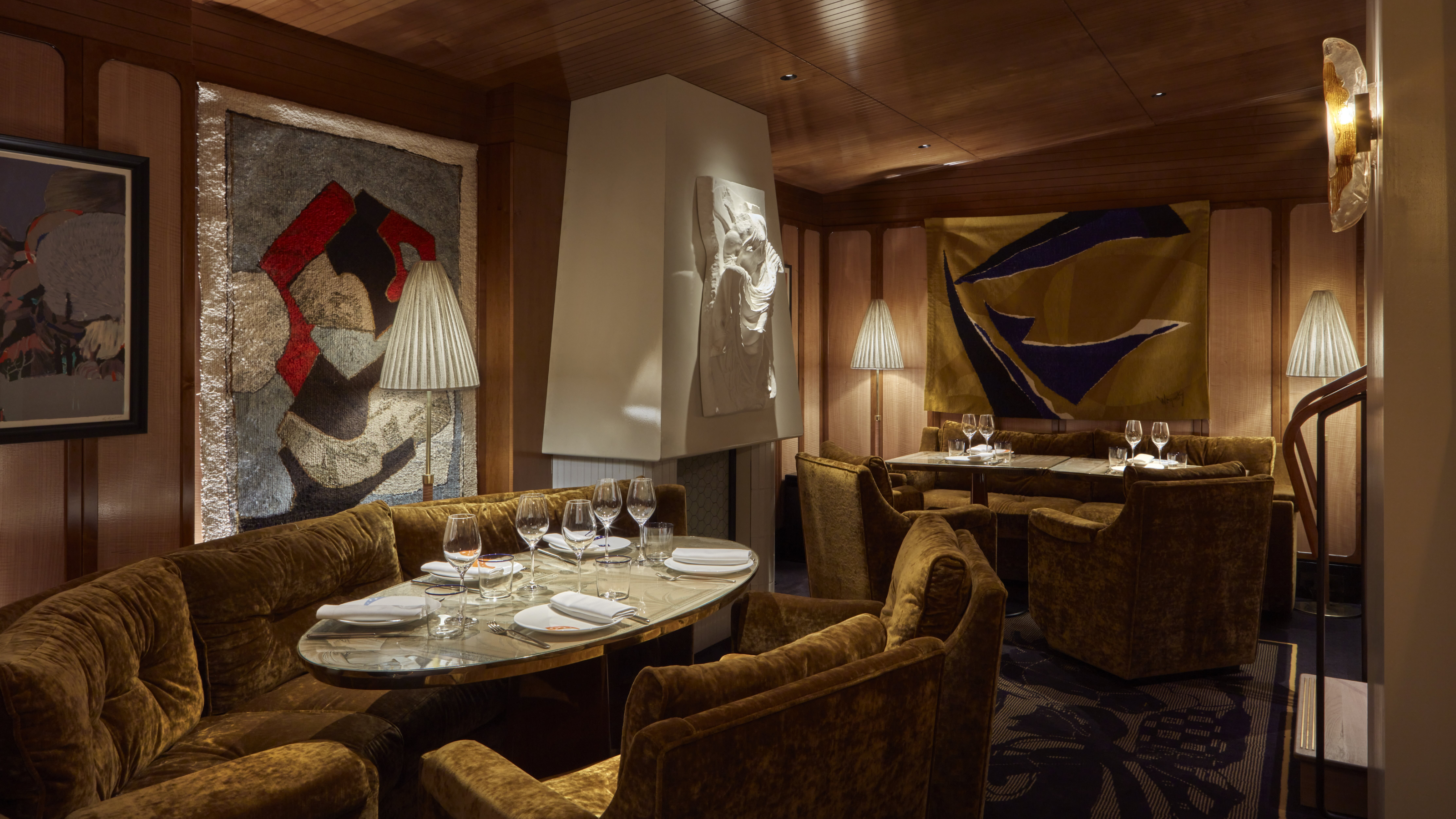 A golden-lit restaurant dining room draped in plush fabrics, velvety banquettes, and colorful artworks, as well as sculptures.