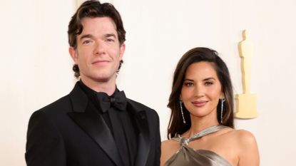 John Mulaney wears an all-black tuxedo while Olivia Munn wears a bronze dress while attending the Academy Awards