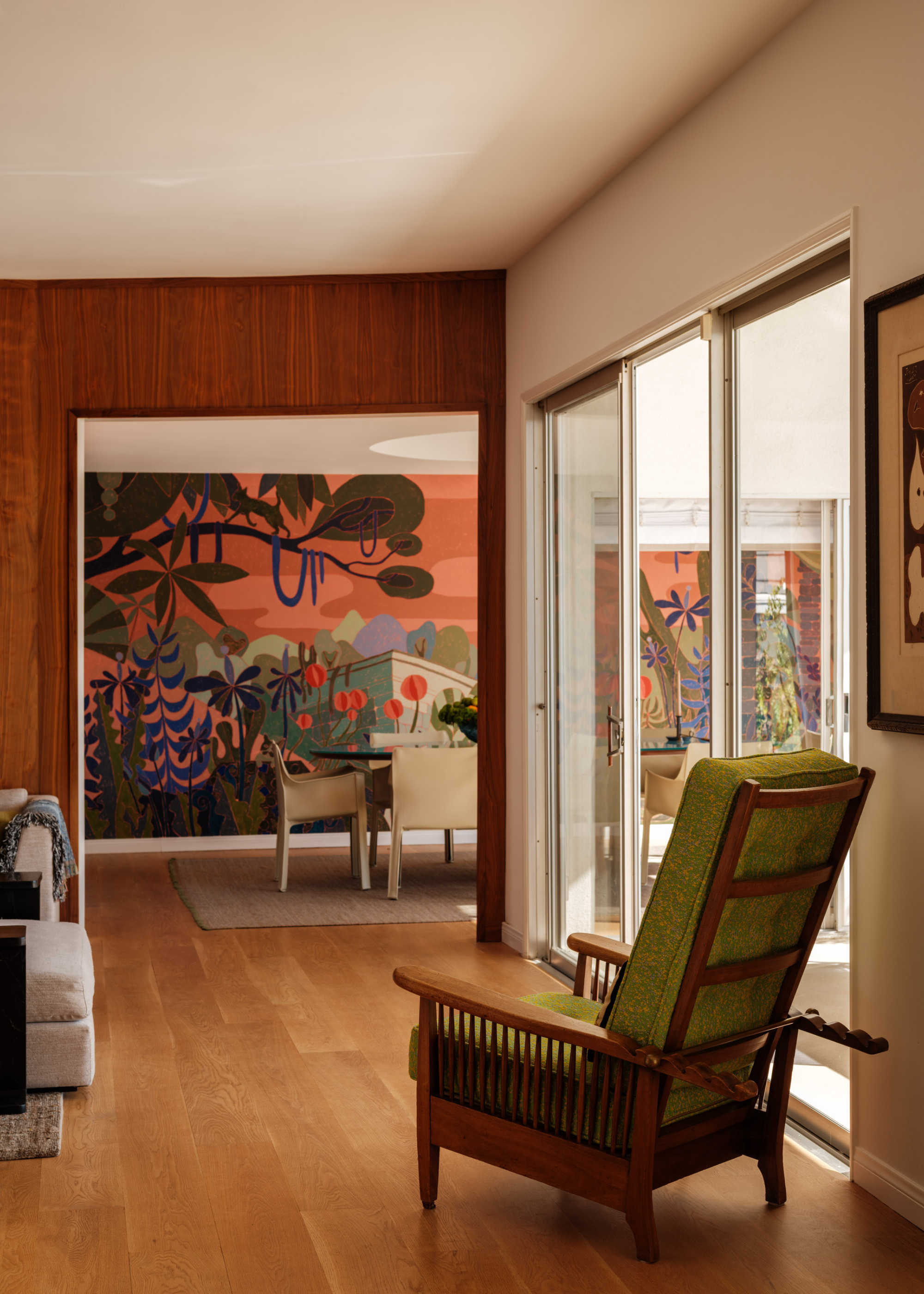A bright living room corridor with sliding doors leading outside, a peak into a dining room with a colorful nature-inspired mural, and a green armchair