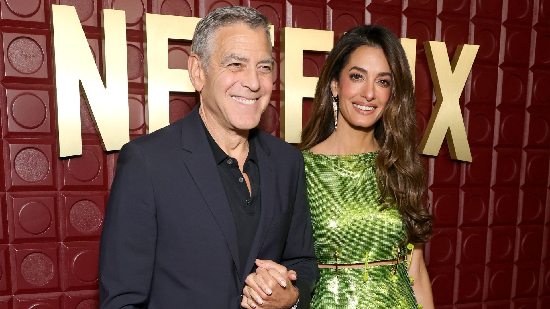 Amal Clooney walks on the red carpet with George Clooney wearing a green Versace dress with gold and vinyl heels.