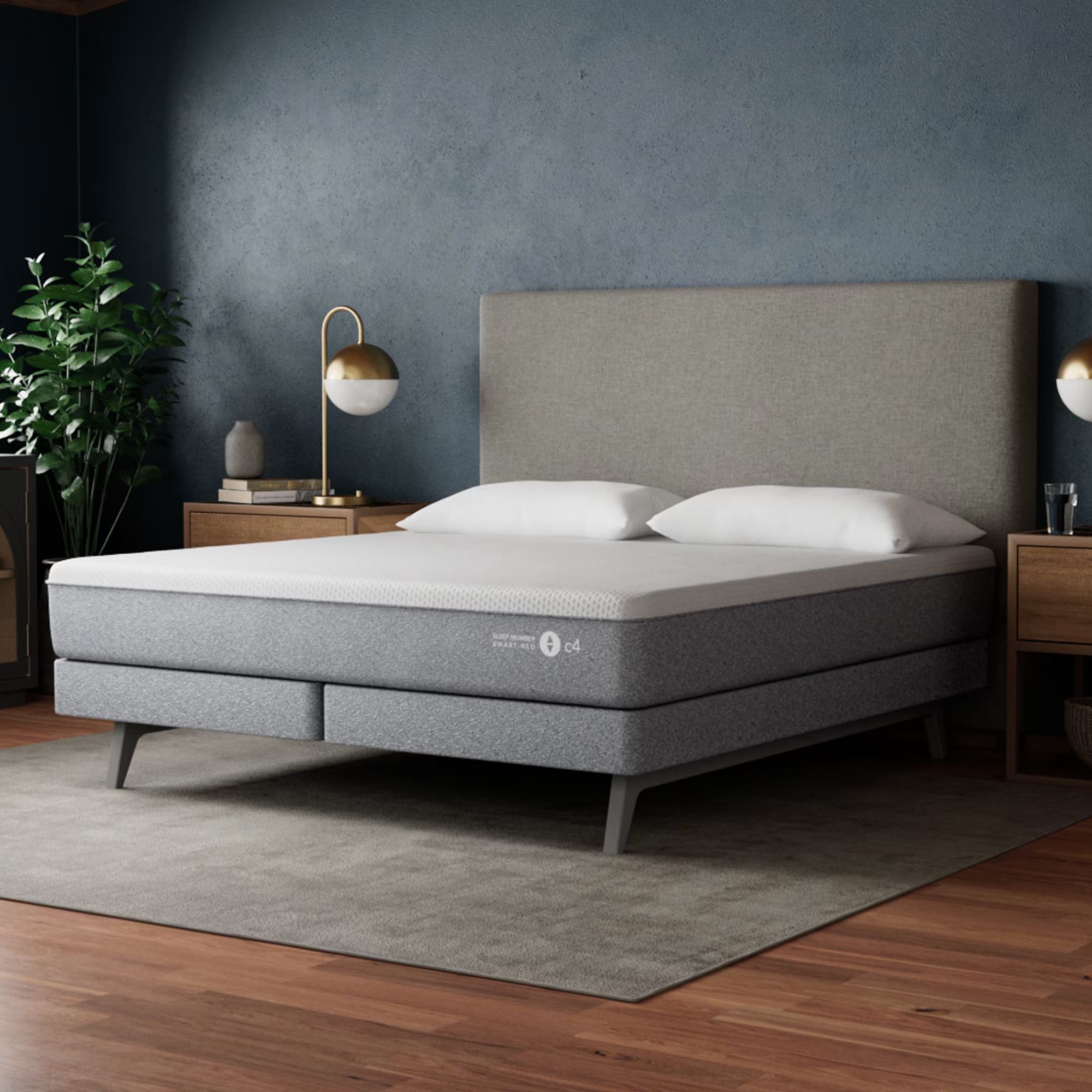Sleep Number c4 mattress shown on a grey bed with large headboard. Beneath the bed, on the floor, is a large grey rug and shined, dark wood flooring, and behind is a dark blue wall. To either side are wooden bedside tables with drawers and matching gold and white lamps. In the far left corner is a large, leafy houseplant.