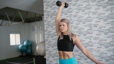 woman standing in a studio setting holding a single dumbbell overhead. she's wearing a black crop top and turquoise leggings. 