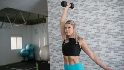 woman standing in a studio setting holding a single dumbbell overhead. she&#039;s wearing a black crop top and turquoise leggings. 