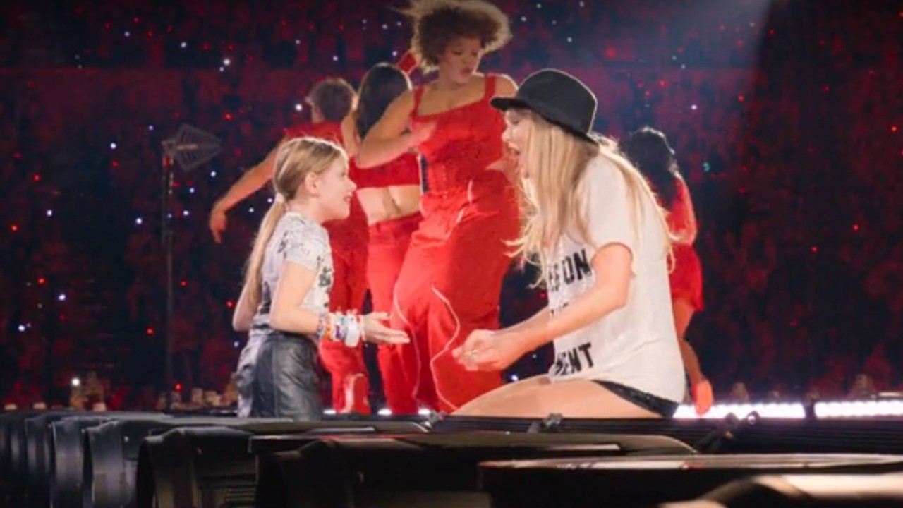 From left to right: A little girl looking at Taylor Swift who is kneeling in front of her while wearing the "22" hat.