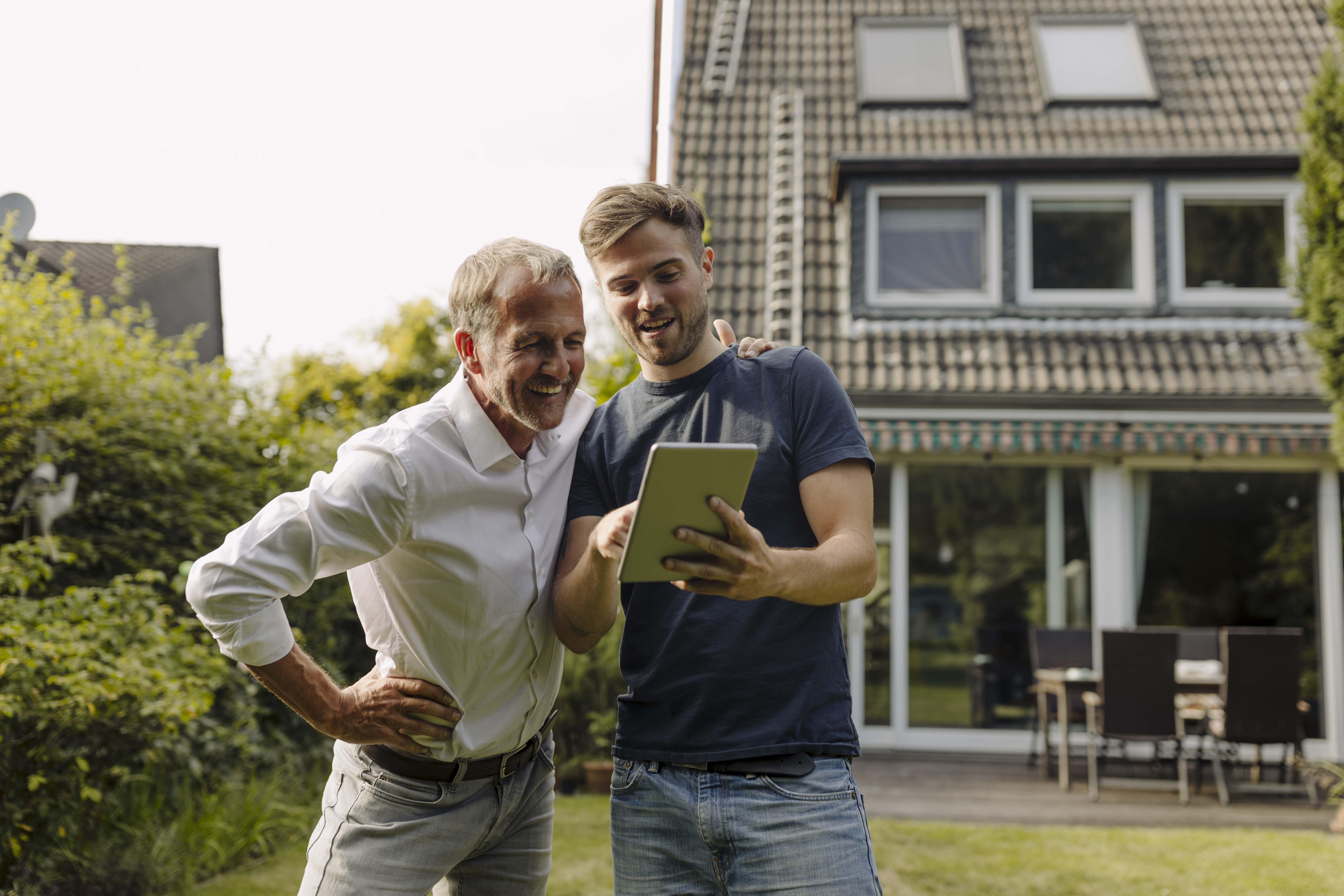 Parent looking to help his adult child buy a house look at tablet as they stand outside a property.