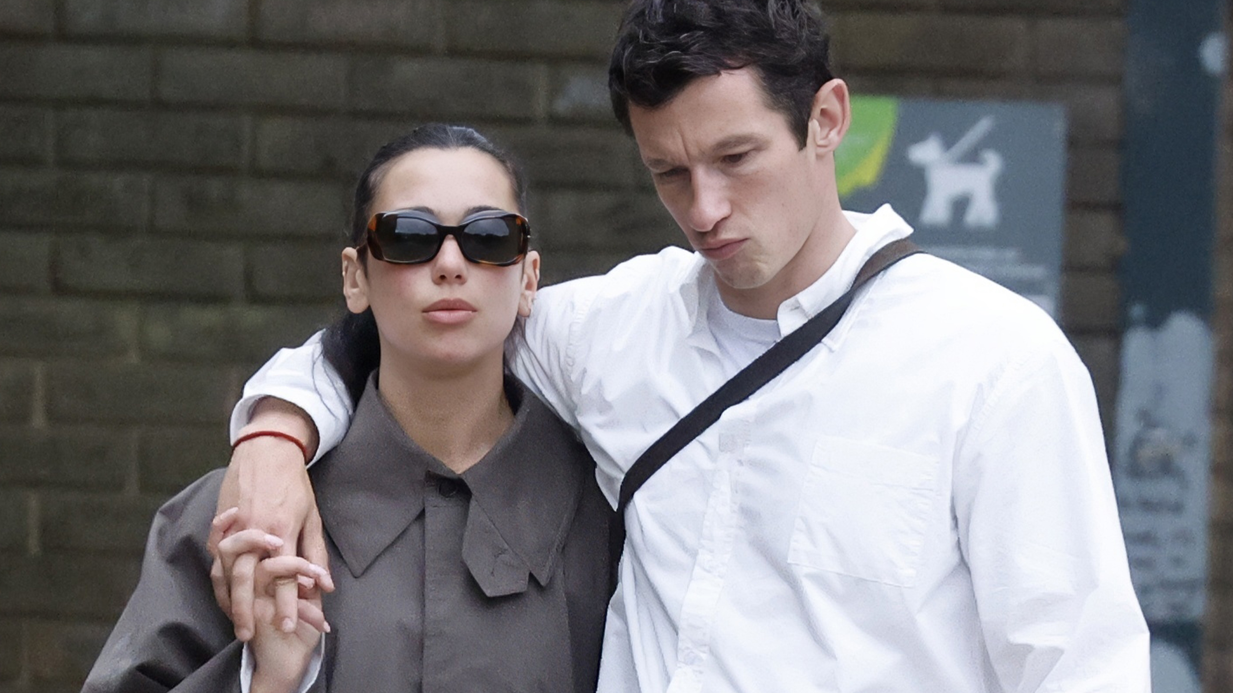 A photo of Dua Lipa and Callum Turner after their London wedding ceremony walking around the city together. Dua is wearing a gray, long trench coat, white jeans, and chestnut Ugg boots.