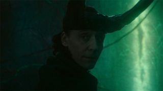Image from the Marvel T.V. show Loki, season 2 episode 6. Close up of Loki's face, the God of Mischief. He is looking back over his shoulder solemnly as he steps towards a green portal. He is wearing a headpiece with two long, black horns on his head.