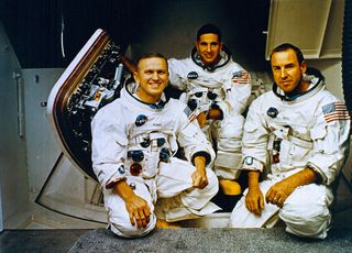 three men in white space suits pose beside a hatch leading into a cone-shaped capsule